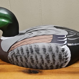 May include: A hand-painted wooden duck decoy. The duck has a green head, black neck, and a white and pink beak. The body is painted with gray, black, and brown feathers. The decoy is sitting on a wooden surface.