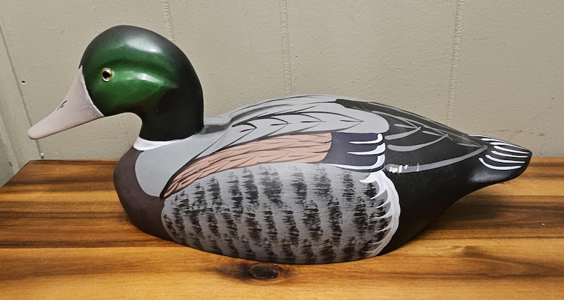 May include: A hand-painted wooden duck decoy. The duck has a green head, black neck, and a white and pink beak. The body is painted with gray, black, and brown feathers. The decoy is sitting on a wooden surface.