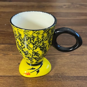Op de afbeelding: Een geel en zwart gespikkelde keramische mok met een zwart handvat en een gele basis. De basis heeft een geschilderd bloemenontwerp. De binnenkant van de mok is wit.