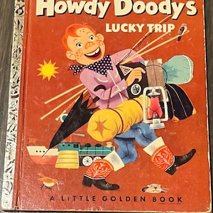 May include: A vintage Little Golden Book featuring Howdy Doody on a lucky trip. The cover shows Howdy Doody, a red-haired puppet with a big smile, carrying a suitcase and other travel items. The book is red with a yellow sun and a blue bird.
