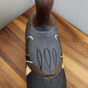 May include: A hand-painted wooden duck decoy. The duck features a brown head with green accents, a dark gray body with black and white details, and a black tail. The decoy is placed on a wooden surface.