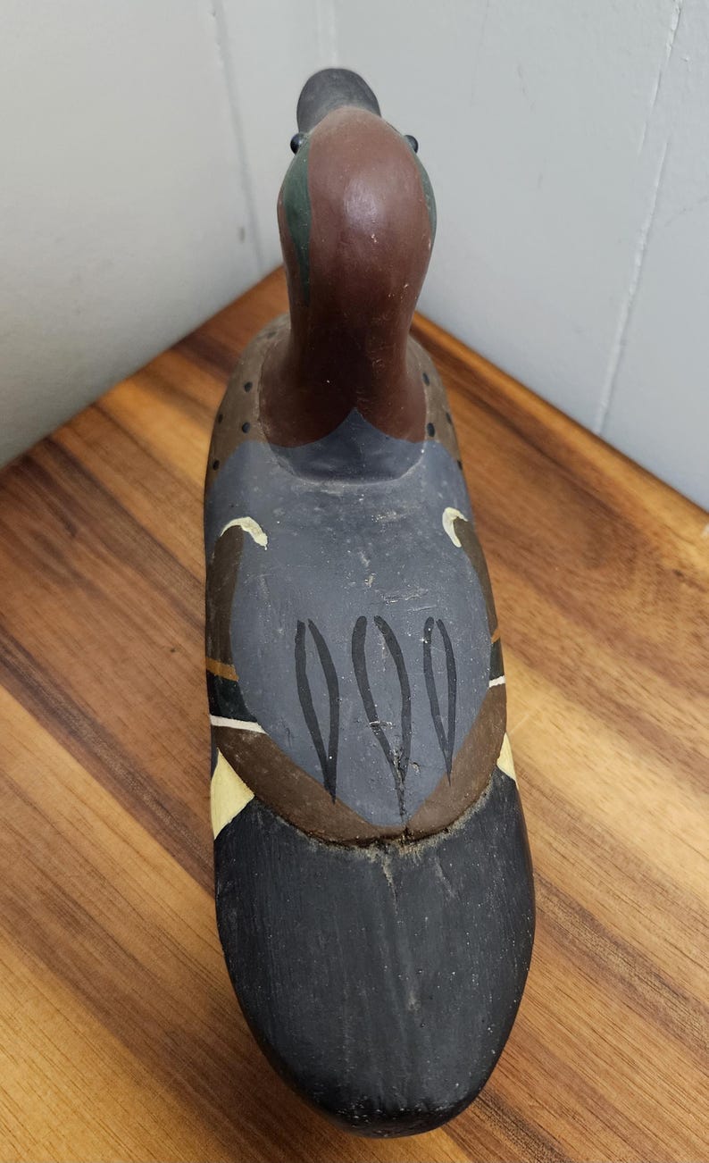 May include: A hand-painted wooden duck decoy. The duck features a brown head with green accents, a dark gray body with black and white details, and a black tail. The decoy is placed on a wooden surface.