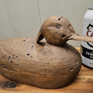 May include: A carved wooden duck decoy with a long beak and detailed feather carvings. The decoy is brown and has several small holes. A can of Canada Dry Zero Sugar Ginger Ale is next to the decoy.