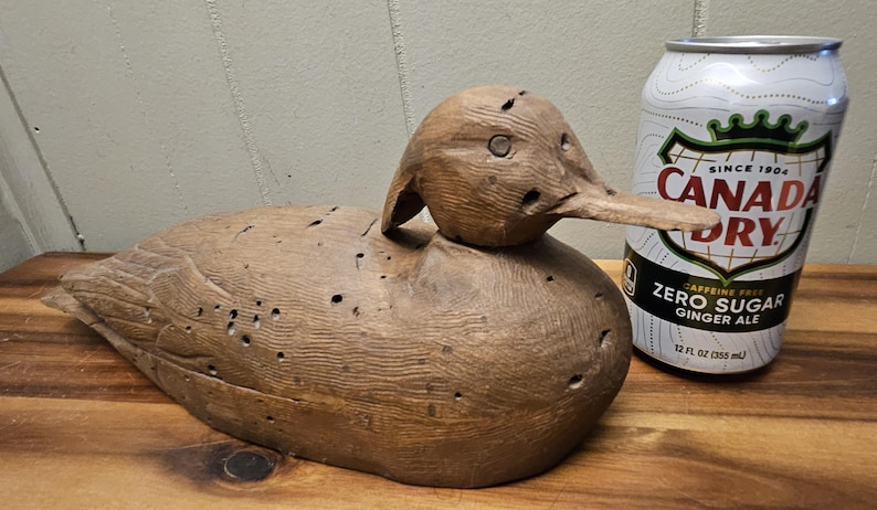 May include: A carved wooden duck decoy with a long beak and detailed feather carvings. The decoy is brown and has several small holes. A can of Canada Dry Zero Sugar Ginger Ale is next to the decoy.