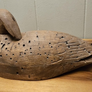 May include: A weathered, wooden duck decoy with a detailed carved design. The duck is a light brown color with visible wood grain and numerous small holes. The decoy is positioned on a wooden surface.