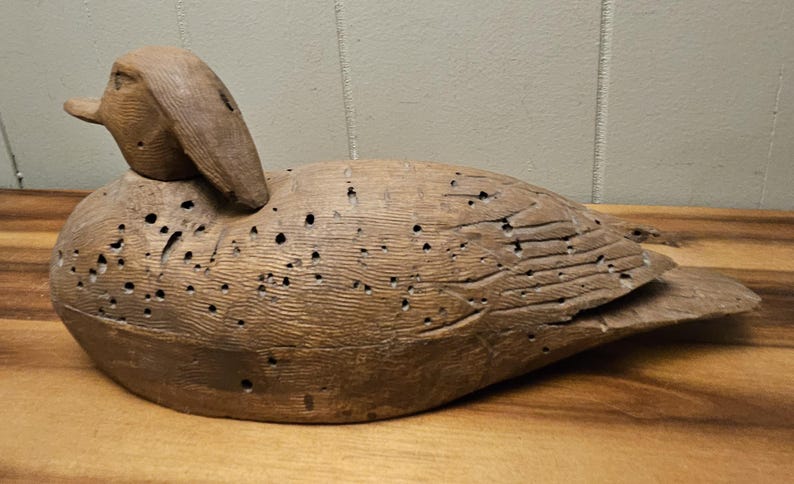 May include: A weathered, wooden duck decoy with a detailed carved design. The duck is a light brown color with visible wood grain and numerous small holes. The decoy is positioned on a wooden surface.