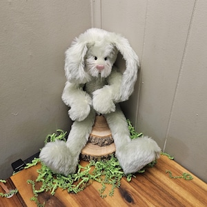 May include: A plush, light green bunny with long, floppy ears and a pink nose. The bunny is seated on a small wooden slice, surrounded by green paper grass, creating a whimsical Easter or spring theme.
