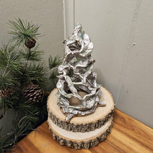 Könnte beinhalten: Eine Keramikskulptur, die einem Baum ähnelt, mit einem strukturierten, geschichteten Design in Weiß und Hellblau. Sie steht auf zwei Holzscheiben mit Rindenkanten. Die Skulptur wird auf einer Holzoberfläche mit Kiefernzweigen und Tannenzapfen im Hintergrund präsentiert.