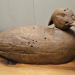 May include: A wooden duck decoy, hand-carved and painted in shades of brown. The duck has a long beak, detailed feather carvings, and several small holes. It rests on a wooden surface.
