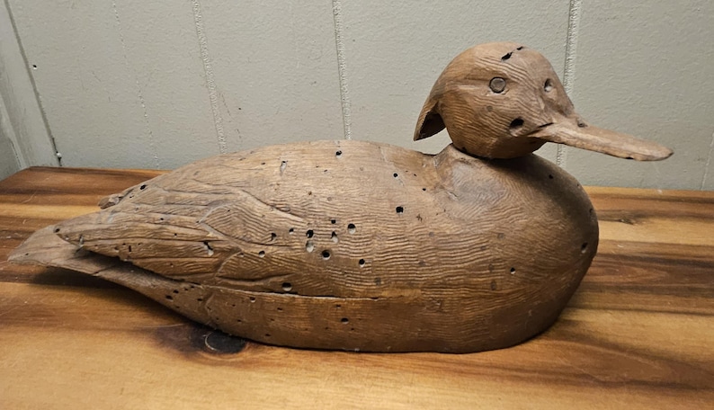 May include: A wooden duck decoy, hand-carved and painted in shades of brown. The duck has a long beak, detailed feather carvings, and several small holes. It rests on a wooden surface.