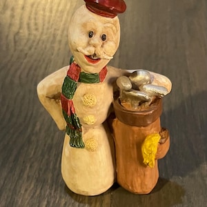 Könnte beinhalten: Eine Schneemann-Figur mit einem roten Barett, einem grün-roten Schal und einem braunen Golfsack mit silbernen Golfschlägern. Der Schneemann hat ein gemaltes Lächeln und einen Schnurrbart.