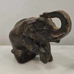 May include: A dark brown, textured elephant figurine depicted in a kneeling position with its trunk curled upward. The sculpture features detailed skin folds and a weathered, matte finish, capturing the animal in a dynamic, grounded pose.