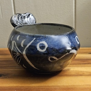 Puede incluir: Un cuenco de cerámica hecho a mano con un esmalte azul oscuro y diseños florales blancos. El cuenco tiene un pequeño asa decorativa y mide aproximadamente 10 cm de diámetro. El cuenco está sobre una superficie de madera.