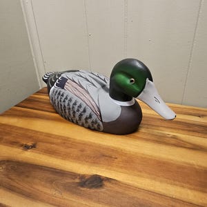 May include: A hand-painted wooden duck decoy. The duck features a green head, gray and brown wings, and a white beak. The decoy is resting on a wooden surface.