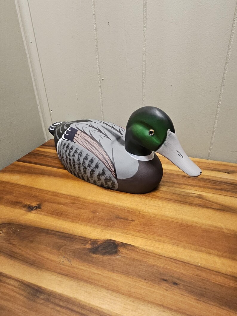 May include: A hand-painted wooden duck decoy. The duck features a green head, gray and brown wings, and a white beak. The decoy is resting on a wooden surface.