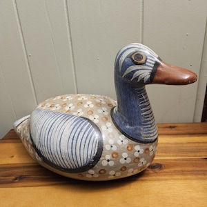 Può includere: Una statuetta di anatra in ceramica dipinta a mano con accenti blu, bianchi e marroni. L'anatra ha un corpo con un motivo bianco e marrone e una testa blu con strisce bianche. Il becco dell'anatra è marrone e i suoi occhi sono gialli.