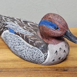 Op de afbeelding: Een handgesneden houten eendenbeeldje. De eend heeft een bruine kop met een blauwe streep, een zwarte snavel en een lichaam met grijs, wit en bruin geschilderde details. De eend rust op een houten oppervlak.