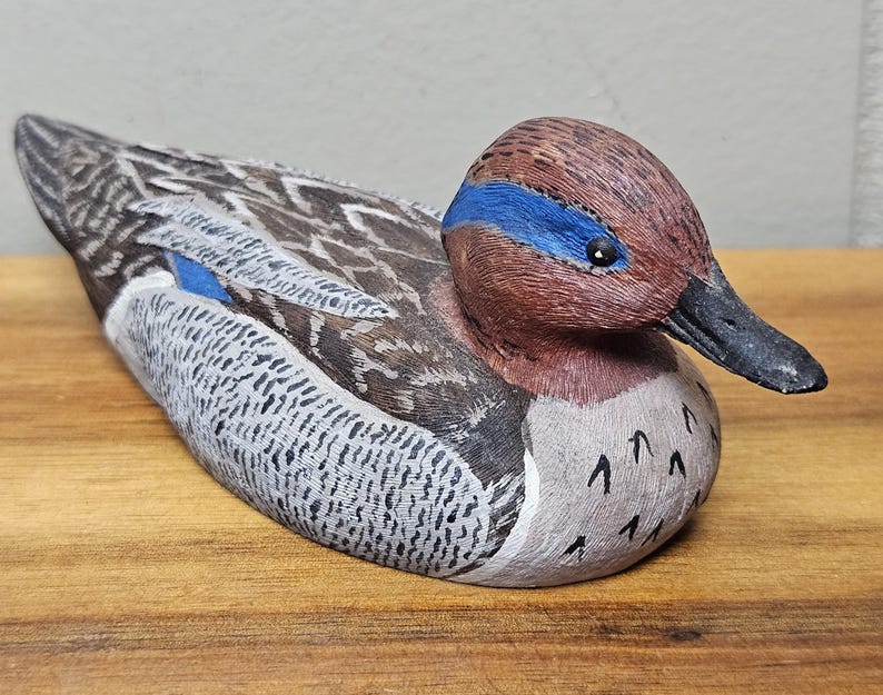 Op de afbeelding: Een handgesneden houten eendenbeeldje. De eend heeft een bruine kop met een blauwe streep, een zwarte snavel en een lichaam met grijs, wit en bruin geschilderde details. De eend rust op een houten oppervlak.