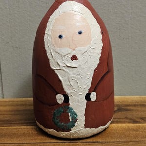 May include: A hand-painted ceramic Santa Claus figurine with a red suit, white beard, and a green wreath in his hand.