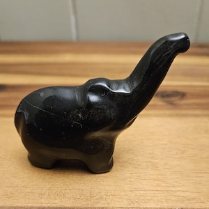 May include: A small, black, carved elephant figurine. The elephant has a raised trunk and is positioned on a wooden surface. The figurine is made of a glossy, dark material, and the details of the elephant's form are clearly visible.