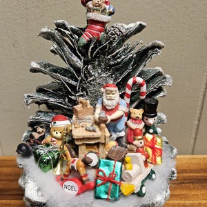 May include: A pine cone Christmas tree decorated with miniature figurines, including Santa Claus, a teddy bear, and a toy train. The tree is also decorated with a candy cane, a stocking, and a variety of other Christmas ornaments. The base of the tree is covered in white cotton batting, and the word "NOEL" is written on the front.
