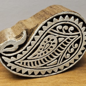 May include: A wooden paisley block with a detailed white design. The block is carved with a central teardrop shape, surrounded by intricate patterns and a sawtooth border. The wood has a natural, aged appearance.