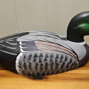 May include: A wooden duck decoy with a green head, black neck, and gray and black body. The duck has a white beak and is resting on a wooden surface. The decoy is painted with detailed feathers.