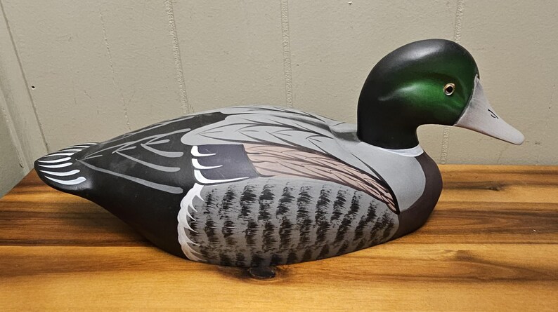 May include: A wooden duck decoy with a green head, black neck, and gray and black body. The duck has a white beak and is resting on a wooden surface. The decoy is painted with detailed feathers.