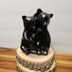 May include: A glossy black ceramic sculpture of two hugging bears. The bears are perched on a rustic, circular wooden base, showcasing a natural bark edge. The sculpture is set against a neutral background.