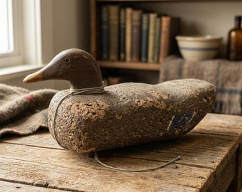 May include: A weathered wooden duck decoy with a dark brown head and a textured, mottled brown body. A thin, light-colored cord is wrapped around the neck, with a loose end hanging down. A small patch of blue paint is visible on the side of the body.