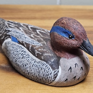 Op de afbeelding: Een handgesneden houten eendenbeeldje. De eend heeft een bruine kop met een blauwe streep, een zwarte snavel en gedetailleerde veermotieven in bruin, grijs en wit. De eend ligt op een houten oppervlak.
