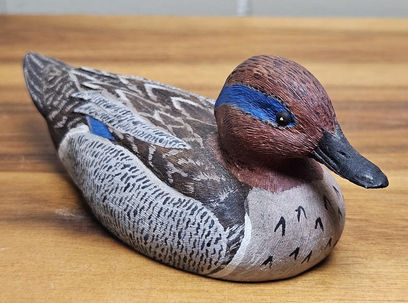 Op de afbeelding: Een handgesneden houten eendenbeeldje. De eend heeft een bruine kop met een blauwe streep, een zwarte snavel en gedetailleerde veermotieven in bruin, grijs en wit. De eend ligt op een houten oppervlak.