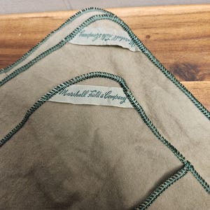 Op de afbeelding: Twee groene vierkanten van stof met een wit label dat op elk is genaaid. Het label op het bovenste vierkant staat "Marshall Field & Company". Het label op het onderste vierkant is gedeeltelijk bedekt, maar lijkt "Marshall Field & Company" te lezen.