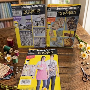 Può includere: Tre buste di cartamodelli Simplicity Sewing Patterns for Dummies, numeri 5750, 4745 e 1140, con marchio giallo e nero. I modelli mostrano vari design per la casa e grembiuli, accompagnati da rocchetti di filo, ritagli di tessuto e forbici.