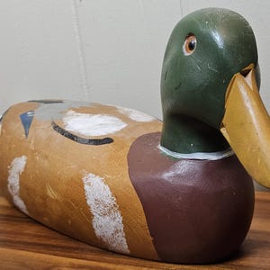 K&ouml;nnte beinhalten: Ein h&ouml;lzerner Entenk&ouml;der mit gr&uuml;nem Kopf, braunem K&ouml;rper und gelbem Schnabel. Die Ente hat wei&szlig;e und schwarze Markierungen auf R&uuml;cken und Seiten. Der K&ouml;der liegt auf einer Holzoberfl&auml;che.