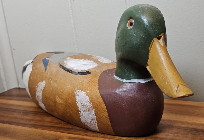K&ouml;nnte beinhalten: Ein h&ouml;lzerner Entenk&ouml;der mit gr&uuml;nem Kopf, braunem K&ouml;rper und gelbem Schnabel. Die Ente hat wei&szlig;e und schwarze Markierungen auf R&uuml;cken und Seiten. Der K&ouml;der liegt auf einer Holzoberfl&auml;che.
