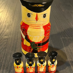 May include: A set of five wooden nutcracker figurines. The largest nutcracker is a tall, cylindrical container with a black hat and a red and yellow painted design. The four smaller nutcrackers are all the same size and have black hats, red and yellow painted designs, and a gold star on their hats.