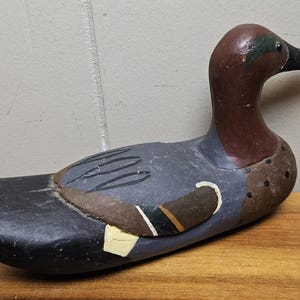 May include: A hand-painted wooden duck decoy. The duck has a brown head and neck, a gray body with black markings, and a black tail. The decoy is resting on a wooden surface.