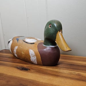 K&ouml;nnte beinhalten: Ein handbemalter Holz-Entenk&ouml;der. Die Ente hat einen gr&uuml;nen Kopf, eine braune Brust und einen gelben Schnabel. Der K&ouml;rper ist in Braun- und Wei&szlig;t&ouml;nen bemalt. Der K&ouml;der liegt auf einer Holzoberfl&auml;che.
