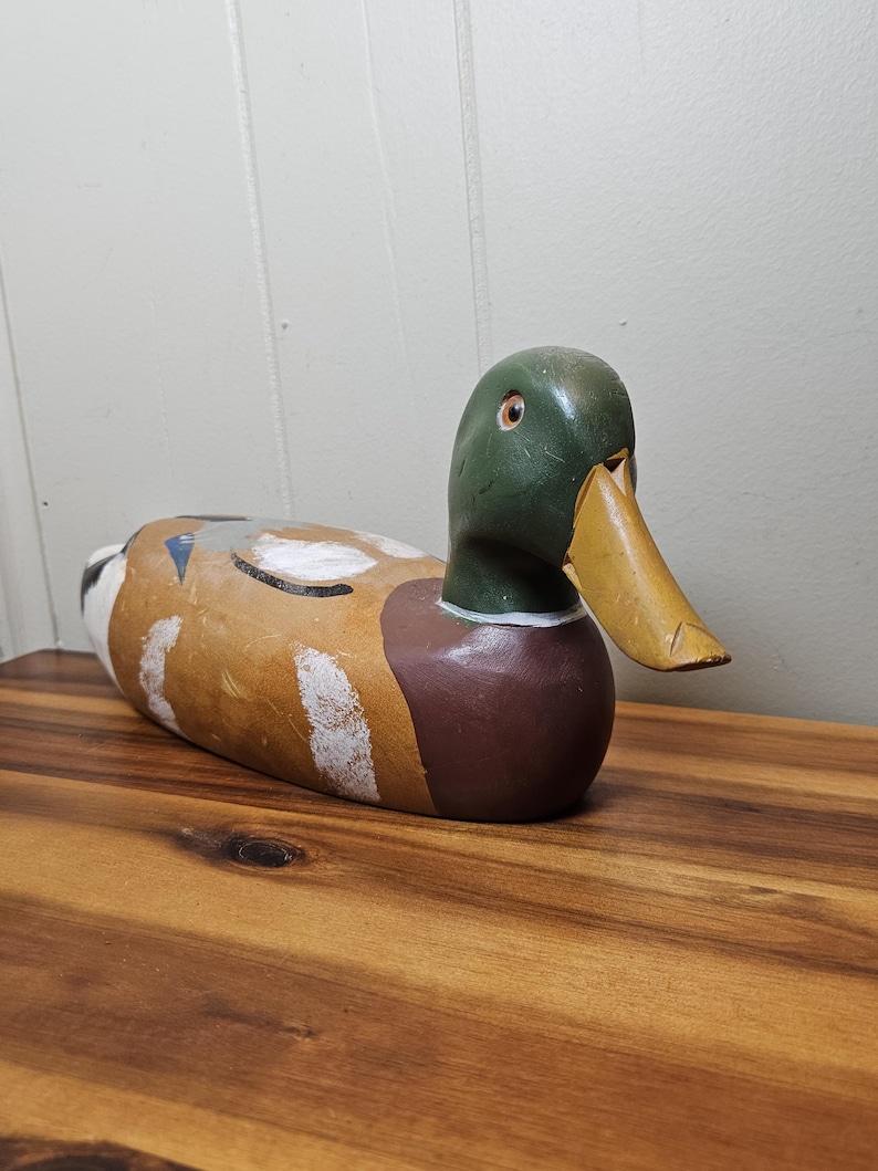 K&ouml;nnte beinhalten: Ein handbemalter Holz-Entenk&ouml;der. Die Ente hat einen gr&uuml;nen Kopf, eine braune Brust und einen gelben Schnabel. Der K&ouml;rper ist in Braun- und Wei&szlig;t&ouml;nen bemalt. Der K&ouml;der liegt auf einer Holzoberfl&auml;che.