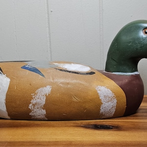 K&ouml;nnte beinhalten: Ein handbemalter Holz-Entenk&ouml;der. Die Ente hat einen gr&uuml;nen Kopf, einen braunen K&ouml;rper und einen gelben Schnabel. Der K&ouml;der ist mit wei&szlig;en, schwarzen und blauen Akzenten bemalt. Er liegt auf einer Holzoberfl&auml;che.