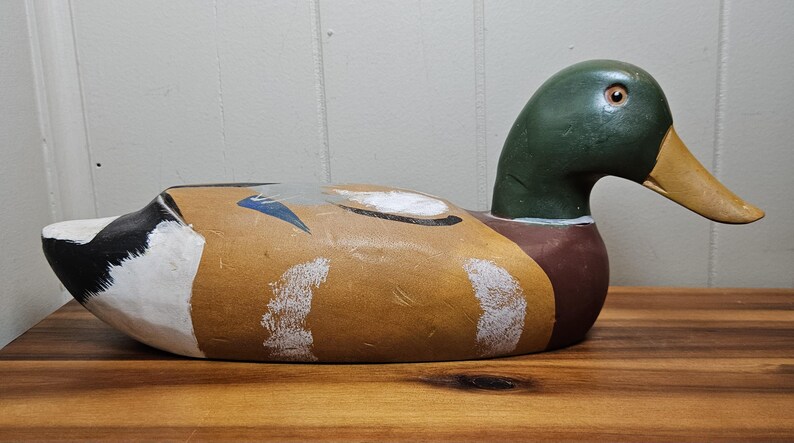 K&ouml;nnte beinhalten: Ein handbemalter Holz-Entenk&ouml;der. Die Ente hat einen gr&uuml;nen Kopf, einen braunen K&ouml;rper und einen gelben Schnabel. Der K&ouml;der ist mit wei&szlig;en, schwarzen und blauen Akzenten bemalt. Er liegt auf einer Holzoberfl&auml;che.