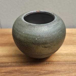 Puede incluir: Un pequeño jarrón redondo de cerámica con un acabado verde metálico texturizado. El jarrón tiene una abertura ancha y se apoya sobre una superficie de madera. El interior es oscuro. Este jarrón decorativo es adecuado para pequeños arreglos florales.