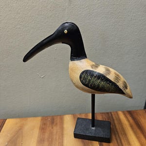 May include: A wooden bird sculpture with a black head and beak, tan body, and black accents. The bird is mounted on a black metal stand with a square base. The sculpture is displayed on a wooden surface.