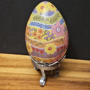 Può includere: Un uovo di Pasqua decorativo in ceramica con un motivo floreale giallo, rosa, arancione e blu. L'uovo è esposto su un supporto in metallo argentato. L'uovo presenta una varietà di fiori e motivi a foglie.