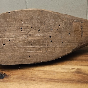 May include: A weathered, wooden duck decoy, showcasing a natural, brown color. The decoy has a rounded body and a pointed tail, with several small holes and visible wood grain. It rests on a wooden surface.