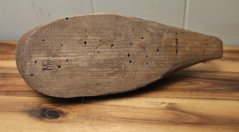 May include: A weathered, wooden duck decoy, showcasing a natural, brown color. The decoy has a rounded body and a pointed tail, with several small holes and visible wood grain. It rests on a wooden surface.