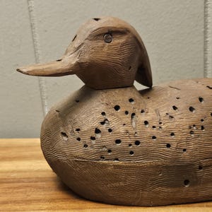 May include: A detailed, hand-carved wooden duck decoy. The duck is brown with a textured surface and numerous small holes. The beak is long and pointed, and the head is slightly tilted. This rustic piece is a classic example of folk art.