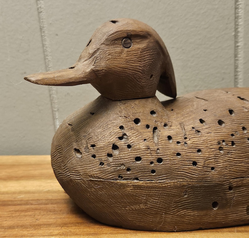 May include: A detailed, hand-carved wooden duck decoy. The duck is brown with a textured surface and numerous small holes. The beak is long and pointed, and the head is slightly tilted. This rustic piece is a classic example of folk art.