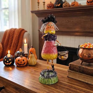 May include: A standing figurine shaped like a piece of candy corn with a yellow, orange, and white body. It wears a black ruffled collar, a black hat, and carries a small owl-shaped basket. The figure stands on a green base with black tinsel.
